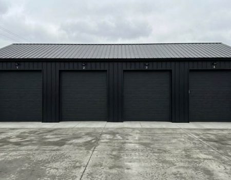 Black 4-bay metal garage with roll-up doors on a concrete slab, modern steel building designed for vehicle storage and workshop use in a commercial setting.