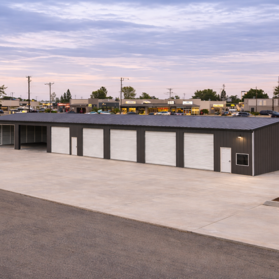 Large charcoal gray commercial metal building with 120-foot length, featuring five white roll-up garage doors and two open workshop bays on a concrete slab at sunset.